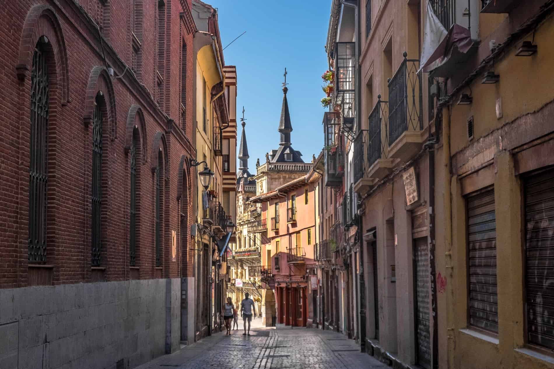 A couple walks up an old street, covered in shade, with illuminated spired buildings in the background. 