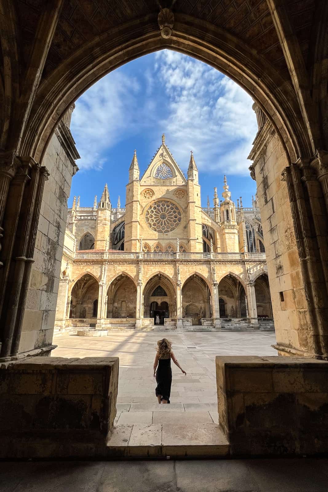 A woman in a black dress walks through a courtyard towards an arcade facade of León Cathedral, Spain.
