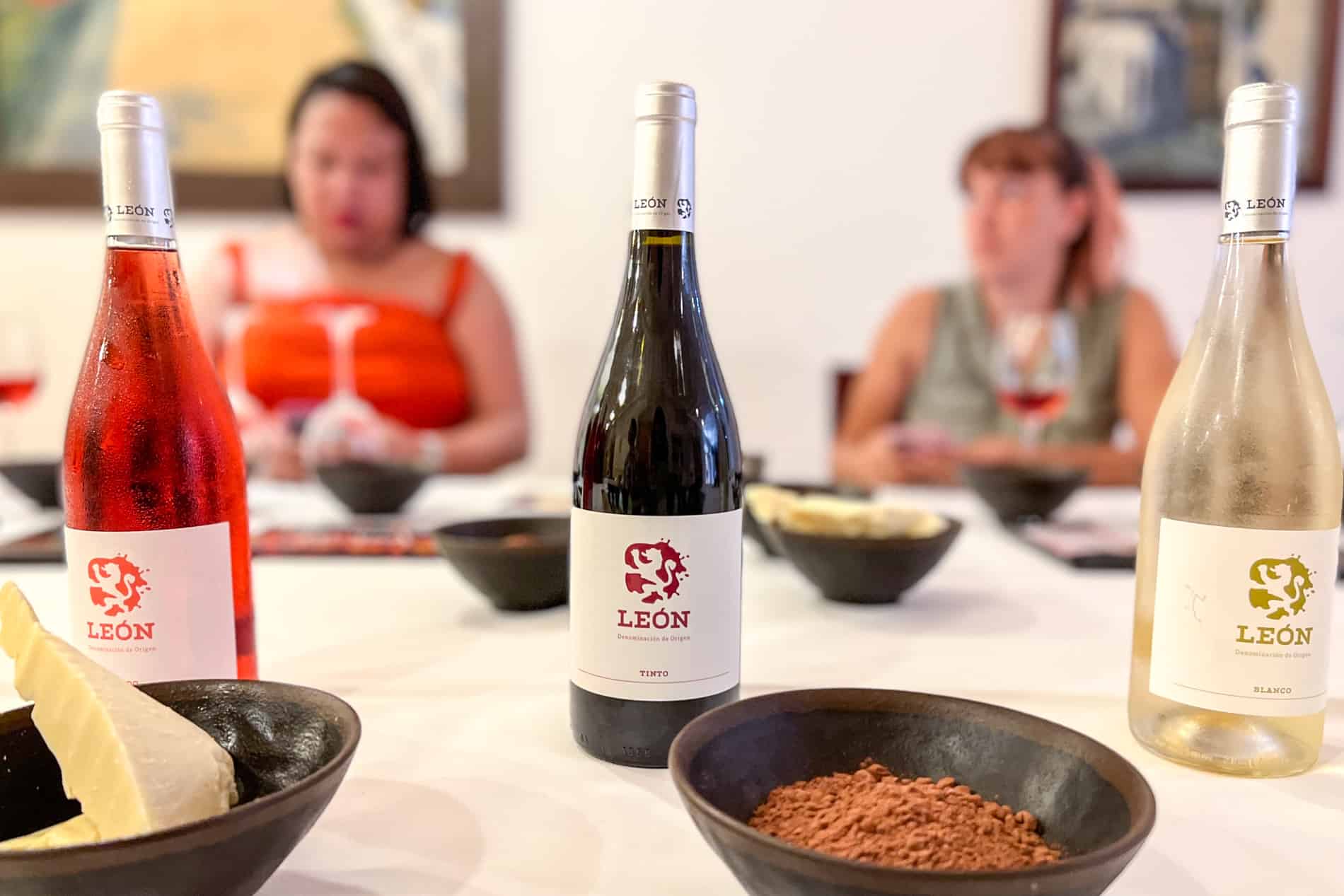 A rose, red and white bottle of wine wlabelled 'León' behind bowls of cacao butter and power and in front of the blurred silhouette of two women sitting at the table. 