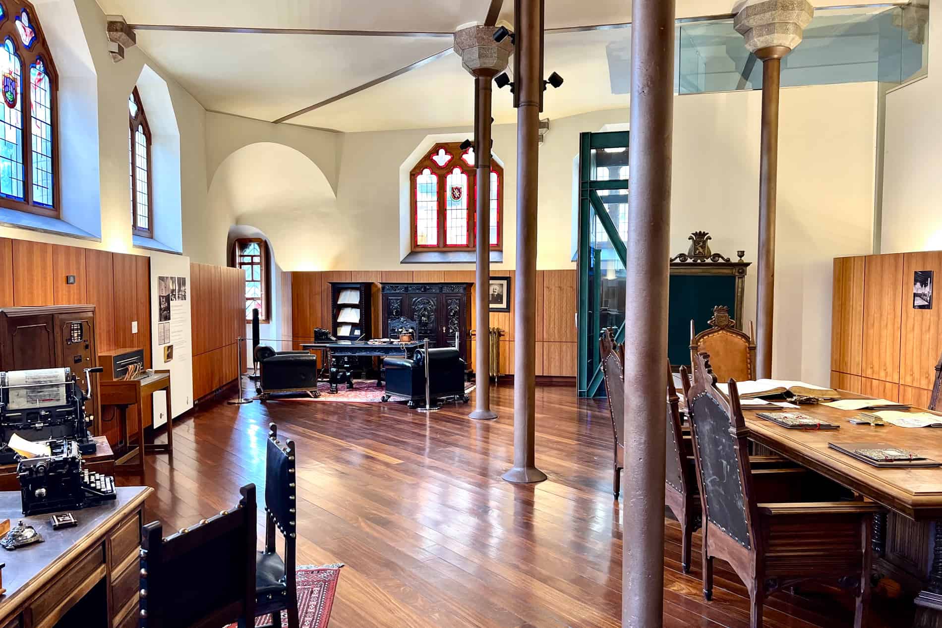 The ground floor of Gaudi's Casa Botines with metal beams and stain glass windows. Three large tables and chairs fill the floor space. 