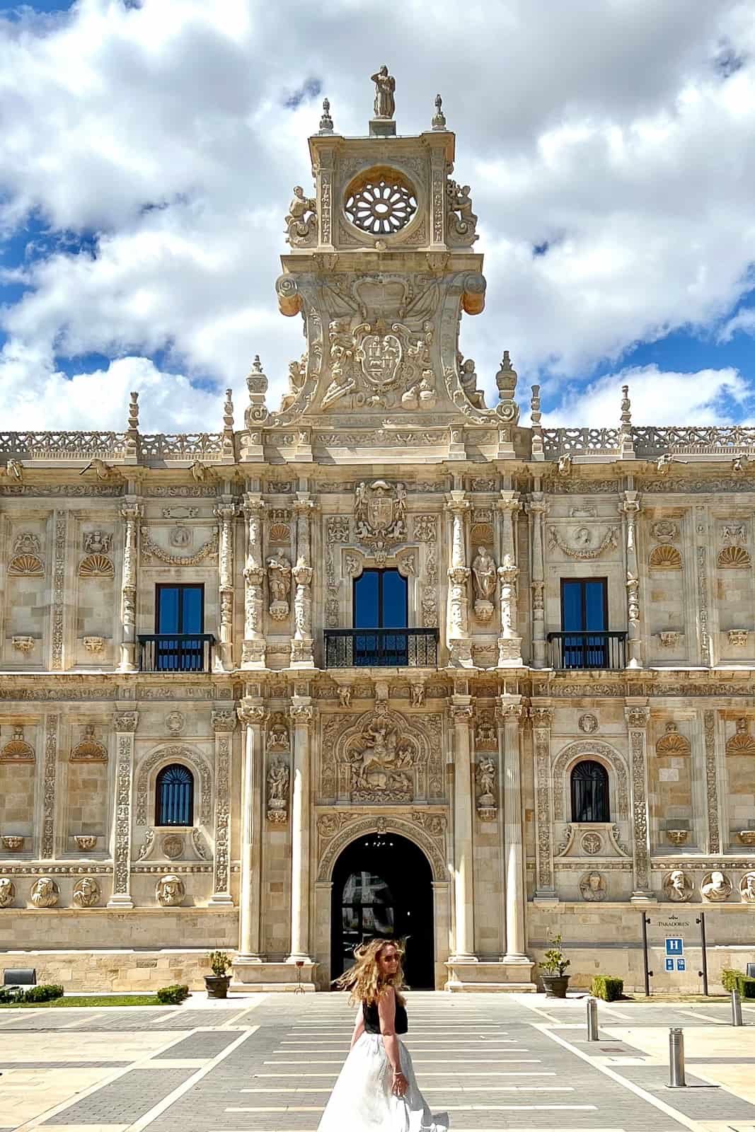 A woman in a black top and white skirt walks towards a Renaissance building in León, Spain and looks back to the camera. 