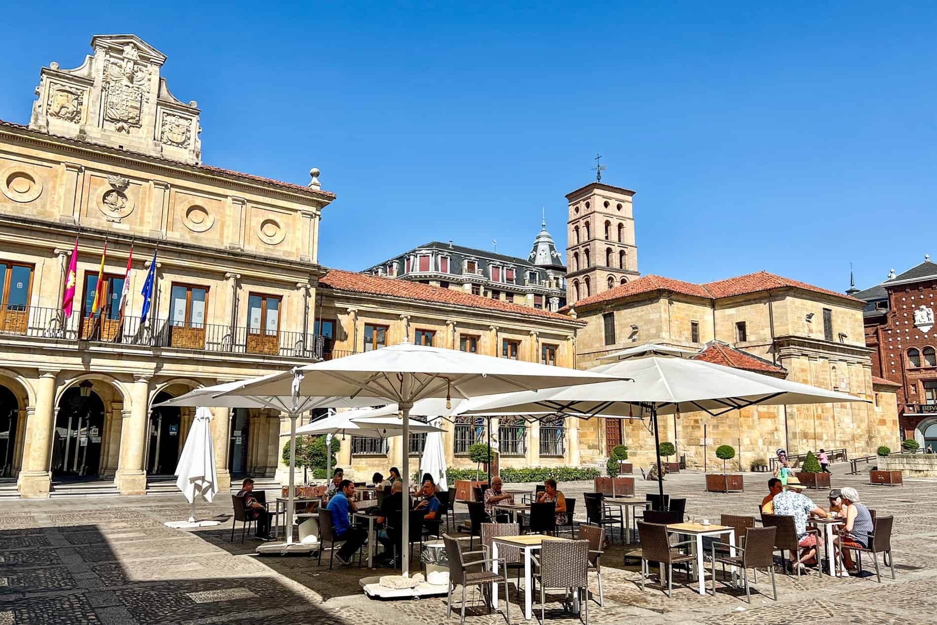 People sitting outside at a cafe under white umbrellas in a public square lined with golden yellow stone buildings. 