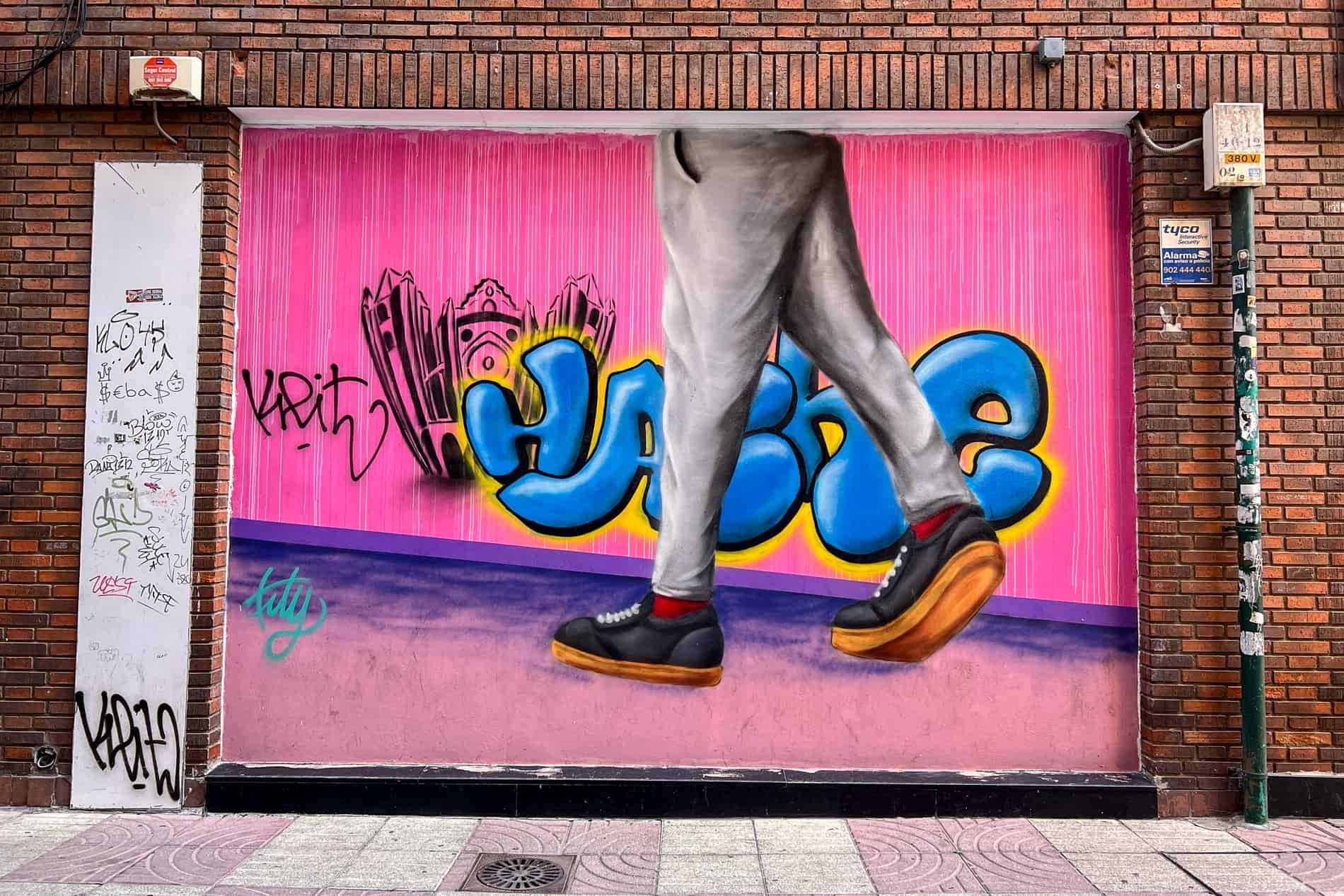 Street art of the lower half of a person walking past a pink wall with blue graffiti lettering. 