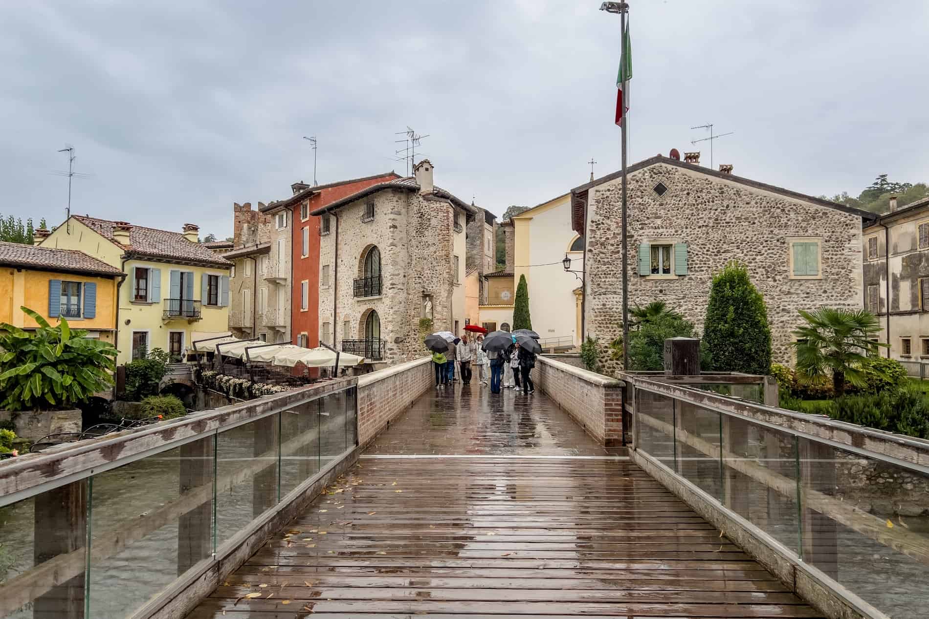 People with umbrellas walk across a wooden bridge towards the stone village houses of Borghetto Sul Mincio.