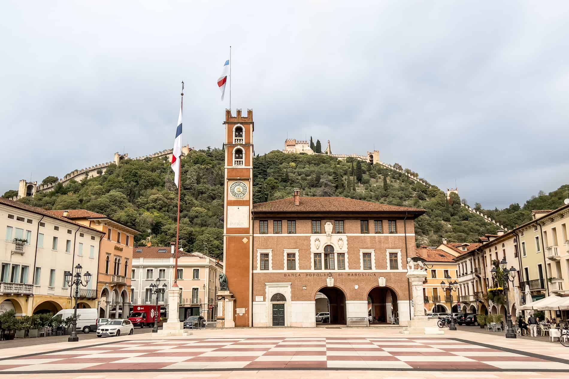 A giant red and white chessboard in a square in Marostica, Italy, backed by a sloping fortress on a hill. 