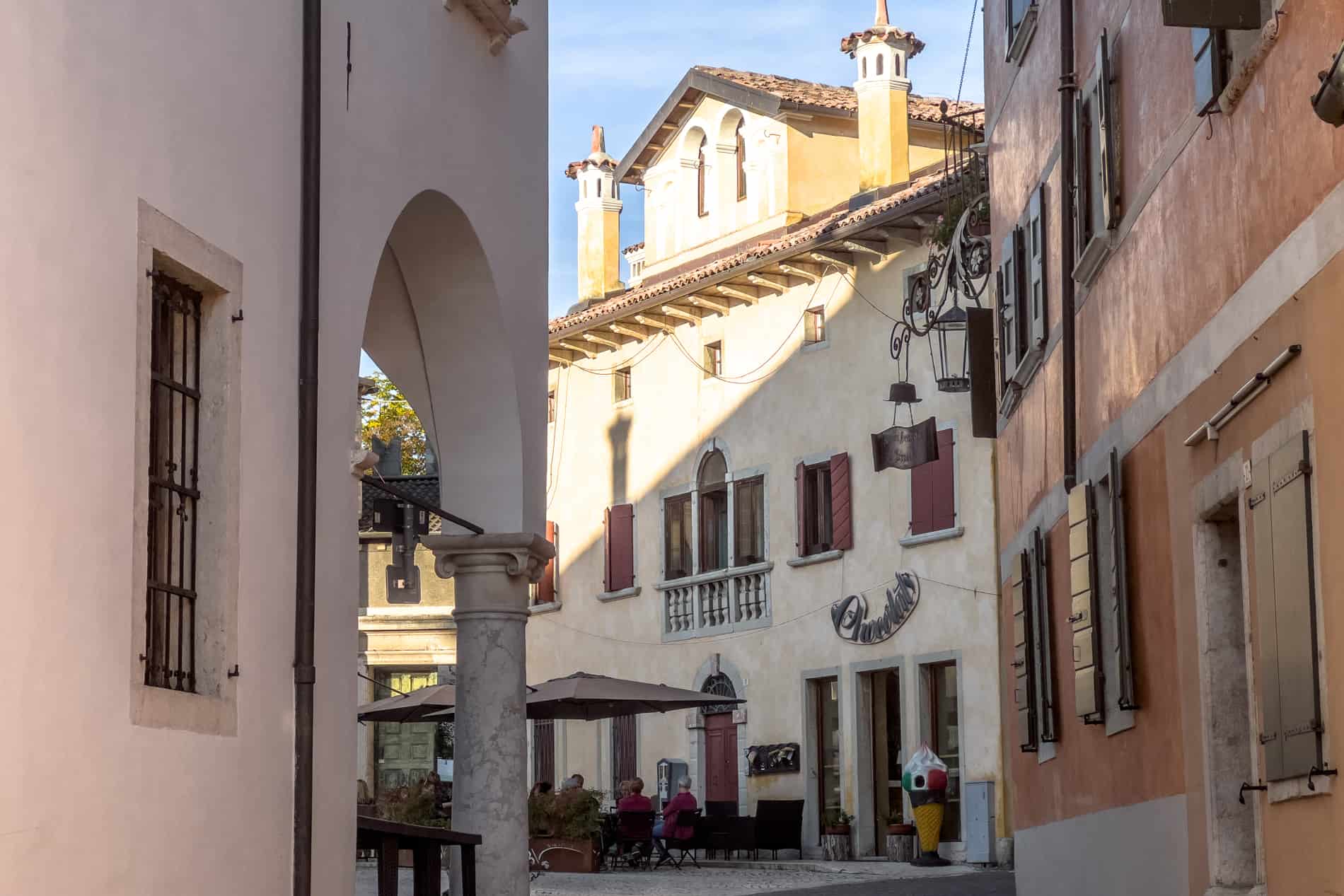 Street scene of three Venetian villa structures converging on a corner, where people are sitting outside under umbrellas. 