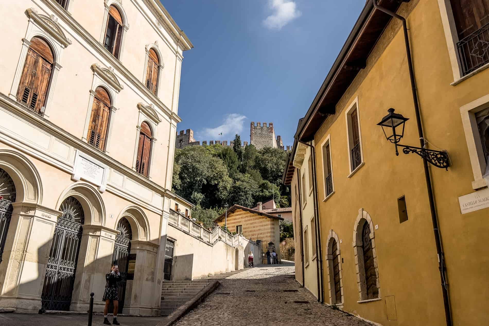 A street in Soave lined with Venetian palazzos, leads to a fortress structure on a forested hilltop. 