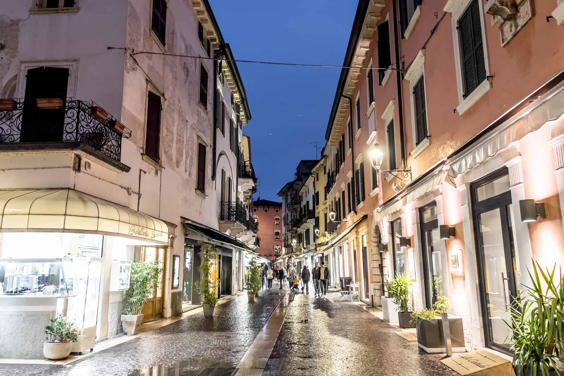 People walking through the narrow stone pastel hued streets of Peschiera del Garda at night, lit up in a yellow glow.