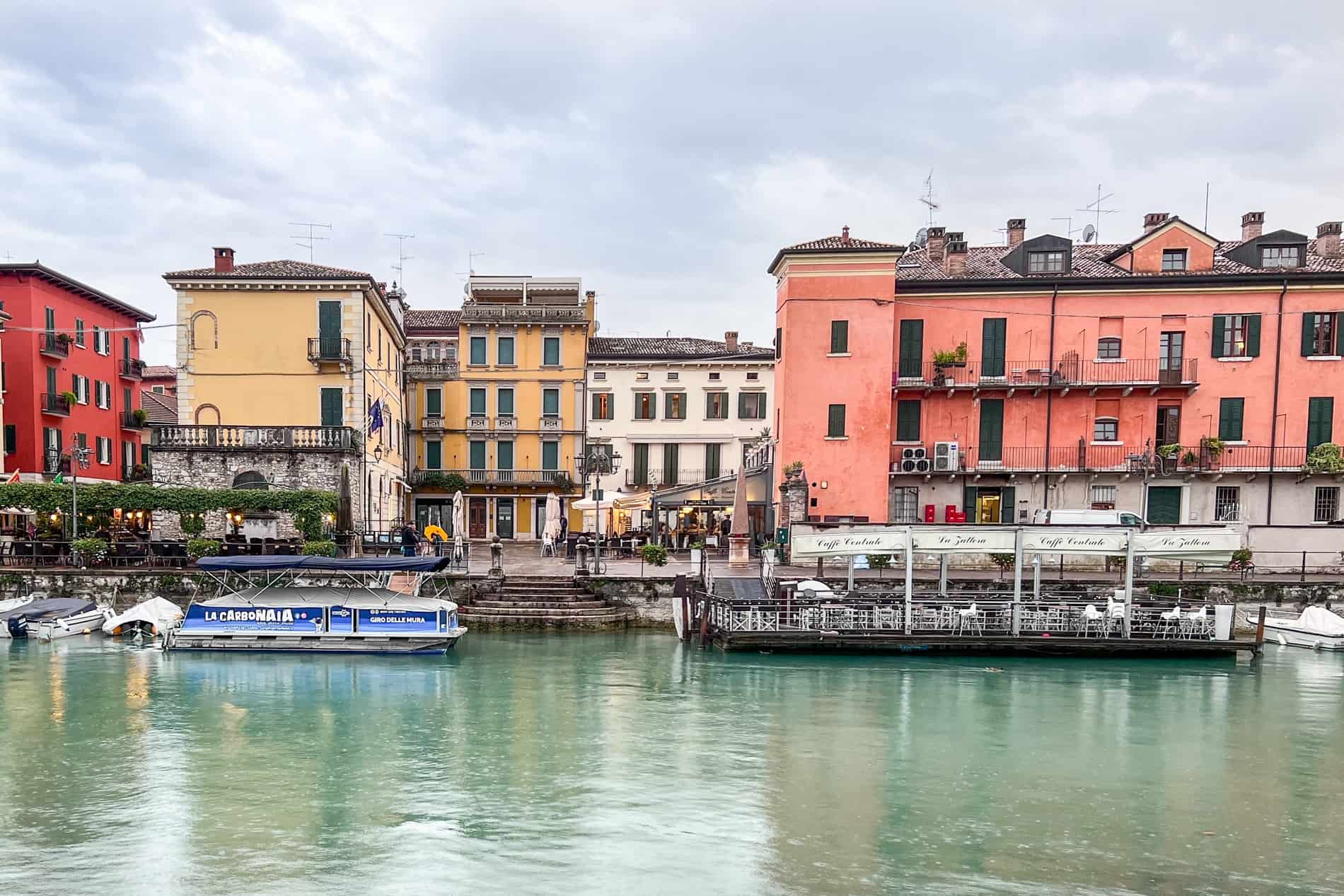 The turquoise canal of Peschiera del Garda lined with red, yellow, and pink buildings of the old town. 