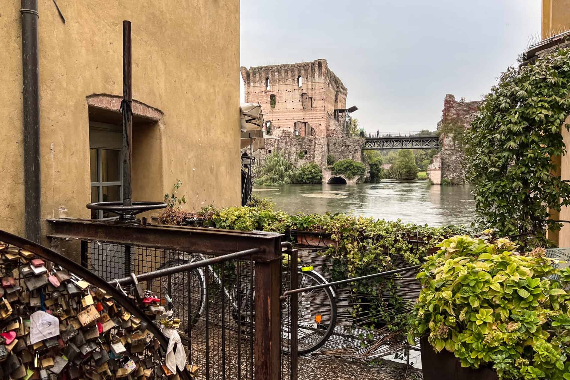 Love locks and a bike on a riverside platform, looking towards a medieval stone tower relic. 