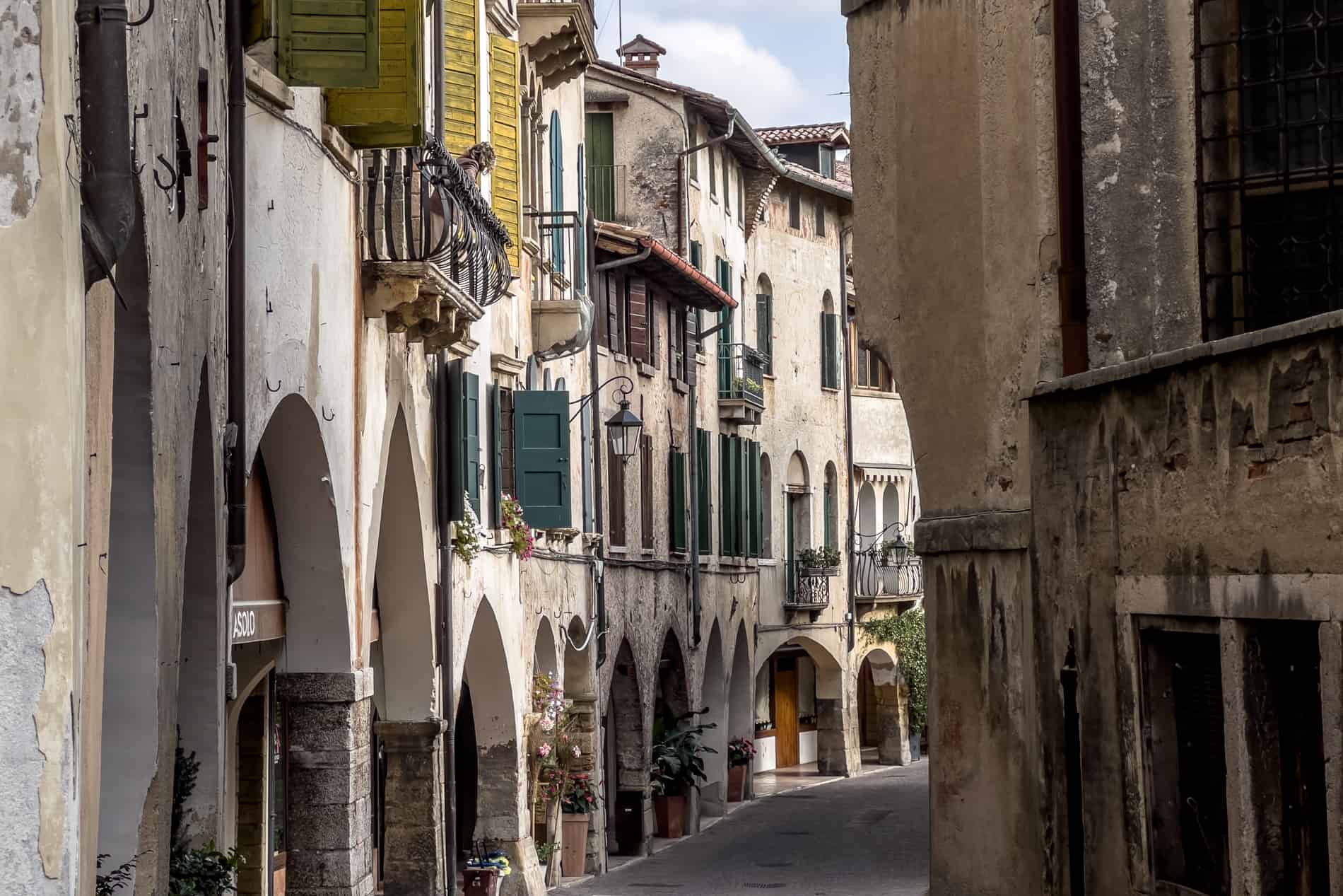 A woman leans over a wooden balcony on a narrow village street with arcades and aged houses.