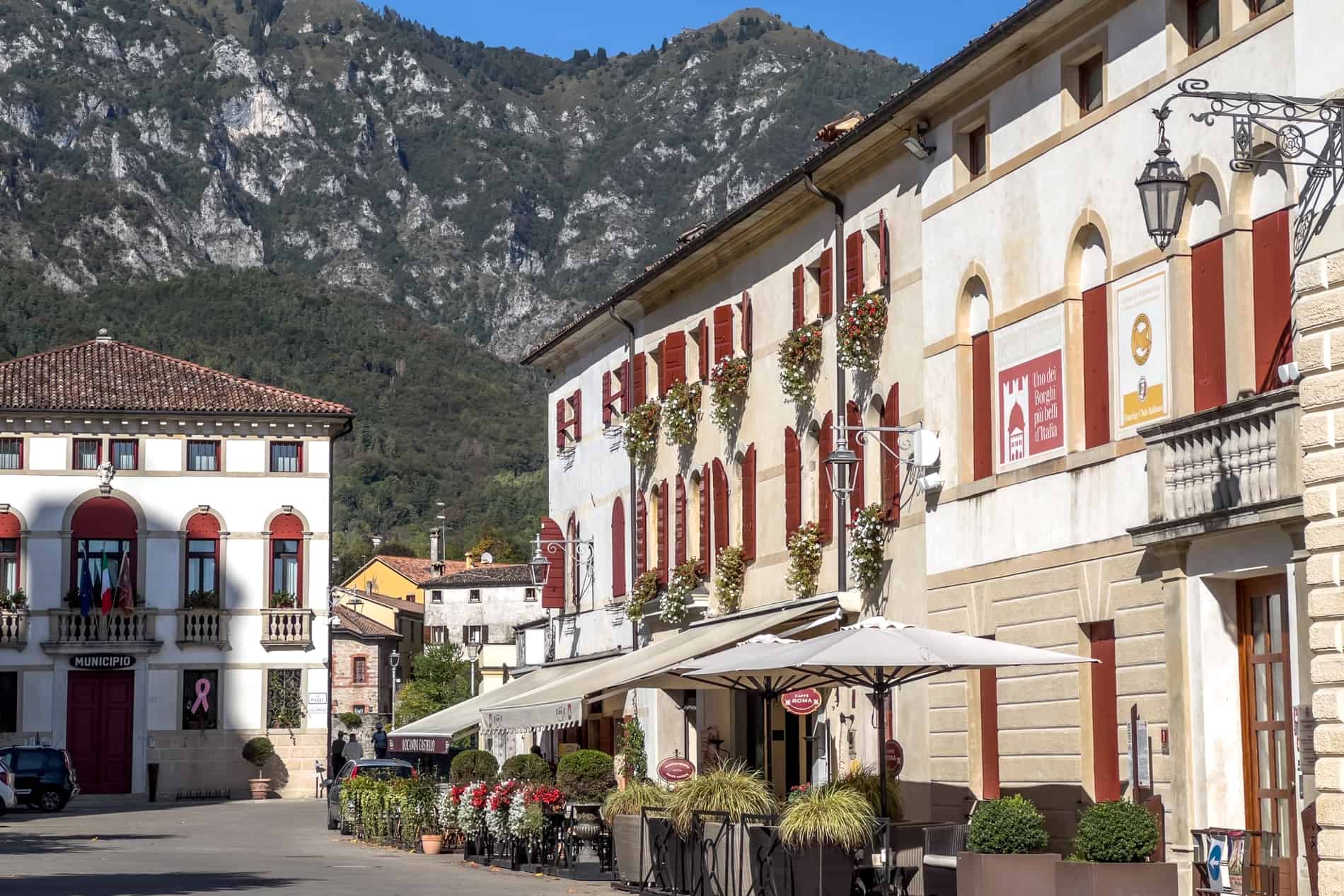 A row of white and beige stone houses with red shutters, in a square backed by low mountains. 