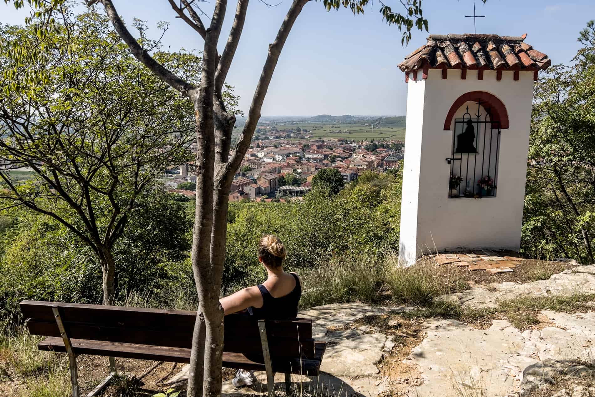 On a hilltop, a woman is sitting on a bench overlooking the cluster of orange buildings of a small village in Italy. 