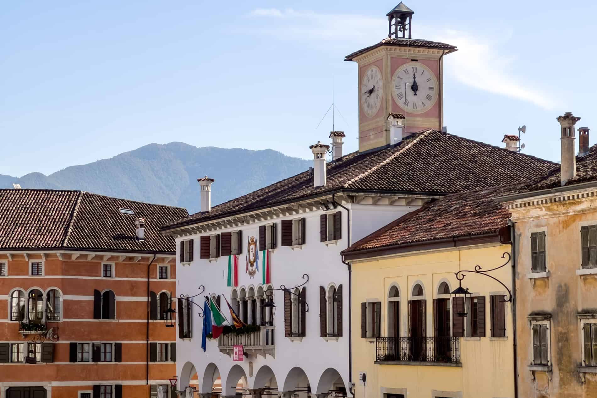 The upper floors of four elegant Venetian palazzos in rust, white (with pink clocktower), yellow and burnt orange colours. 