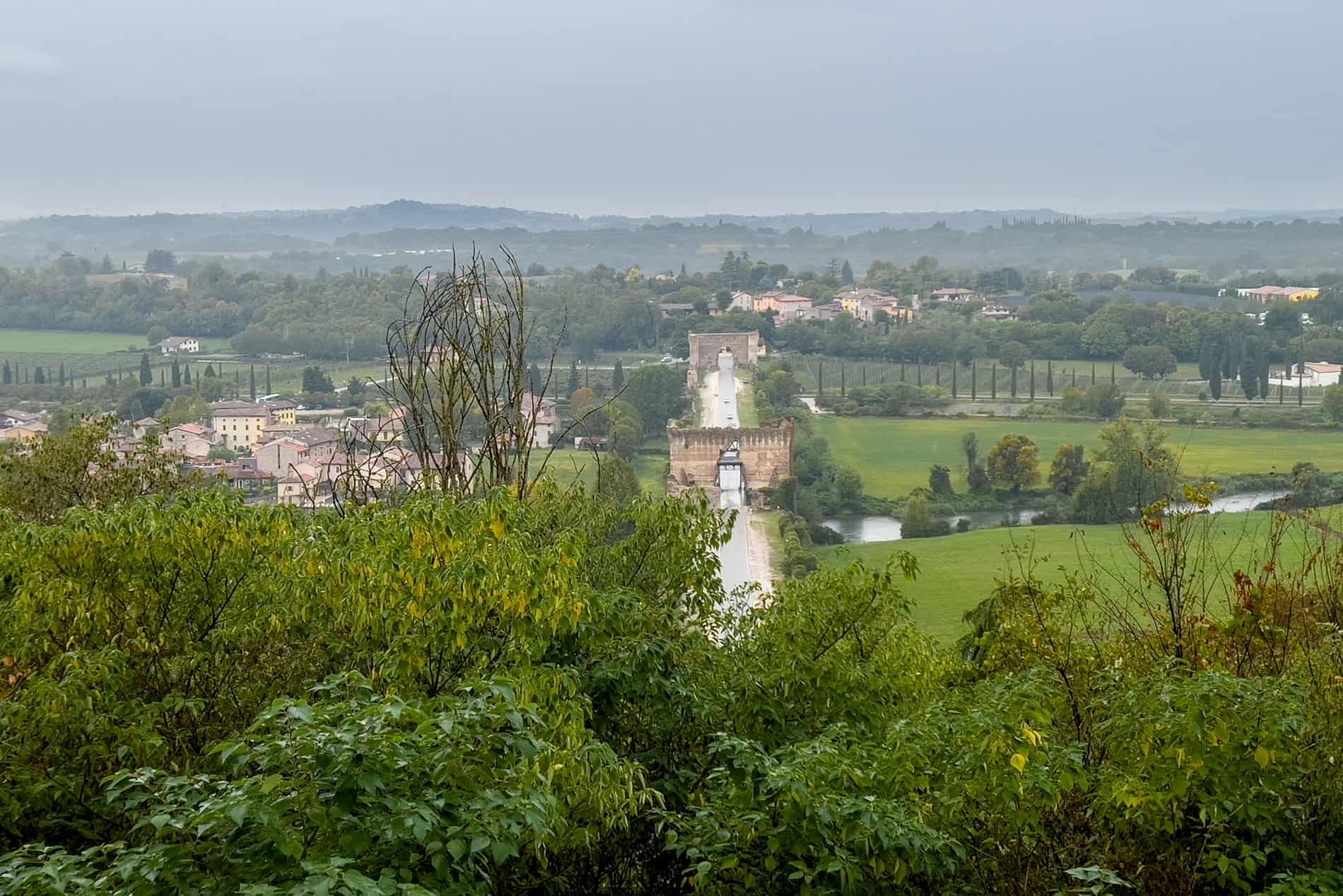 An elevated view over an ancient river bridge structure in Borghetto Sul Mincio, surrounded by green land and trees. 