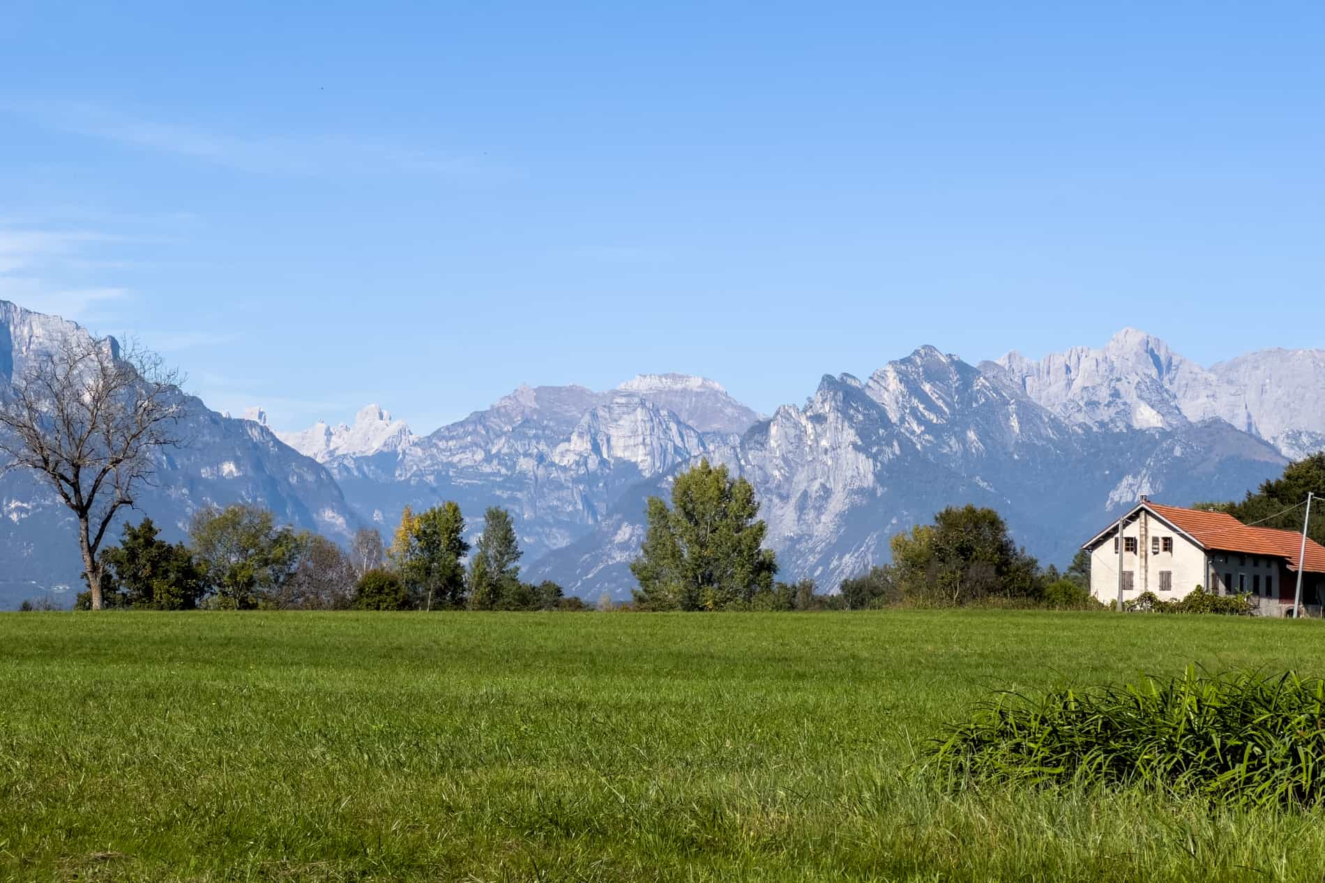 A white farmhouse with orange roof on a wide green field, backed by the Dolomites mountains in the village of Mel, Italy. 