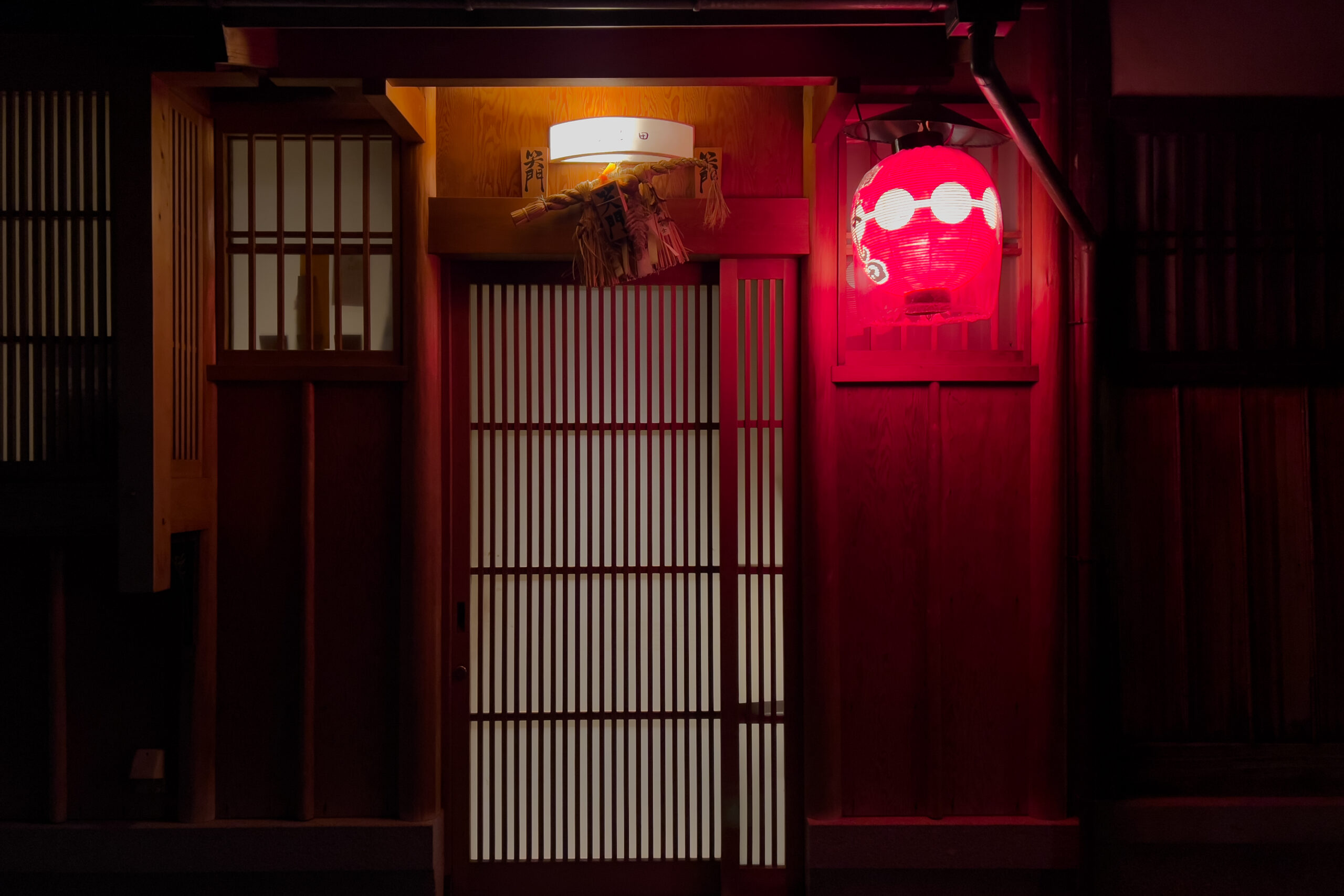 A traditional Japanese machiya entrance in Kyoto at night, featuring a sliding wooden latticed door and a glowing red paper lantern.