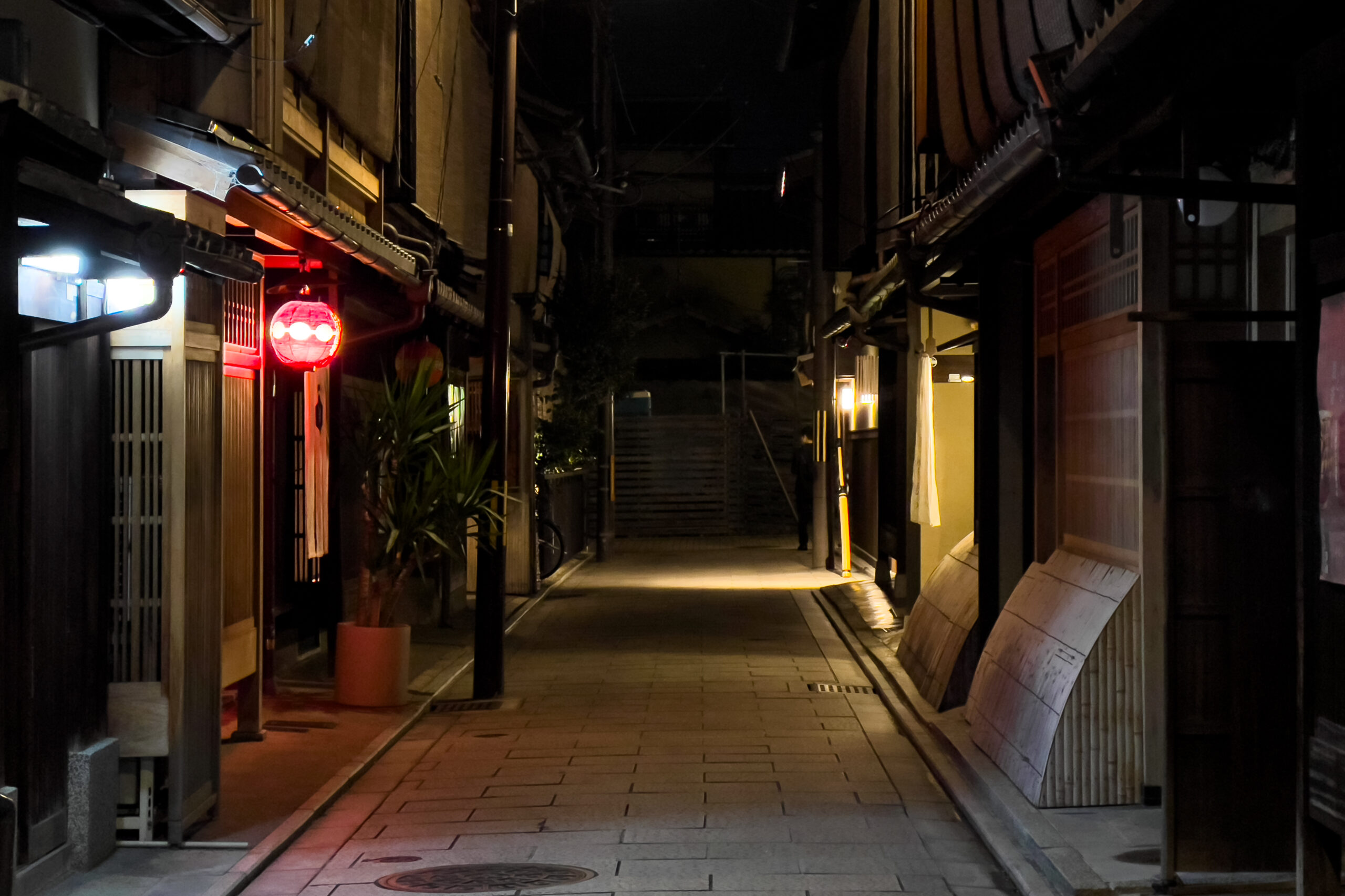 Night view of a narrow, cobblestone alleyway in Gion, Kyoto, featuring traditional wooden machiya houses and a glowing red paper lantern.