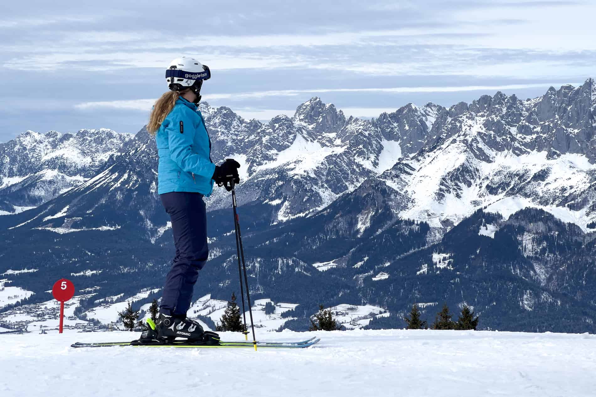 Woman in a light blue ski jacket and dark blue ski pants looks out to a mountain vista. 