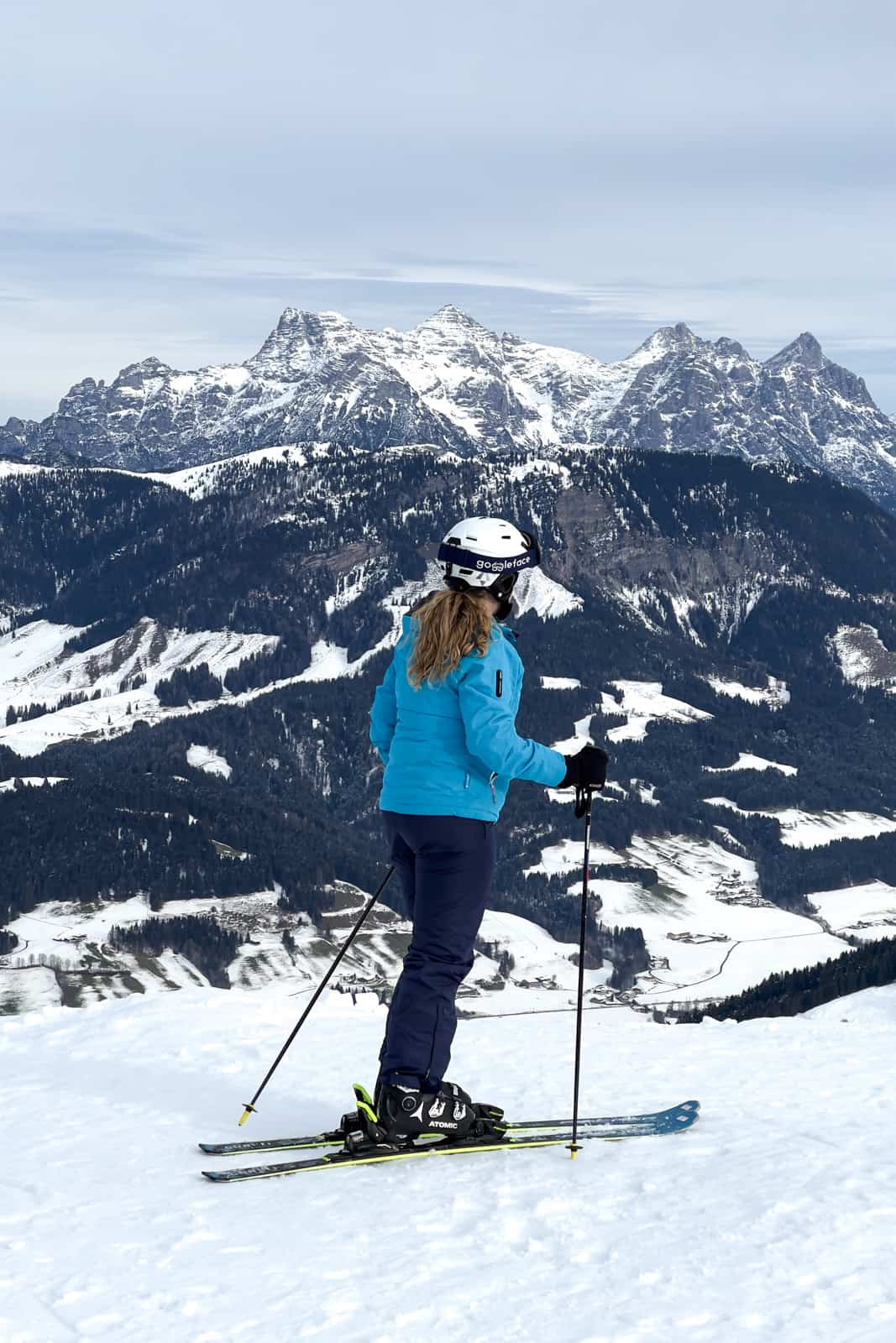 A woman on skis stops to look at the mountain range in the distance, behind the valley below. 