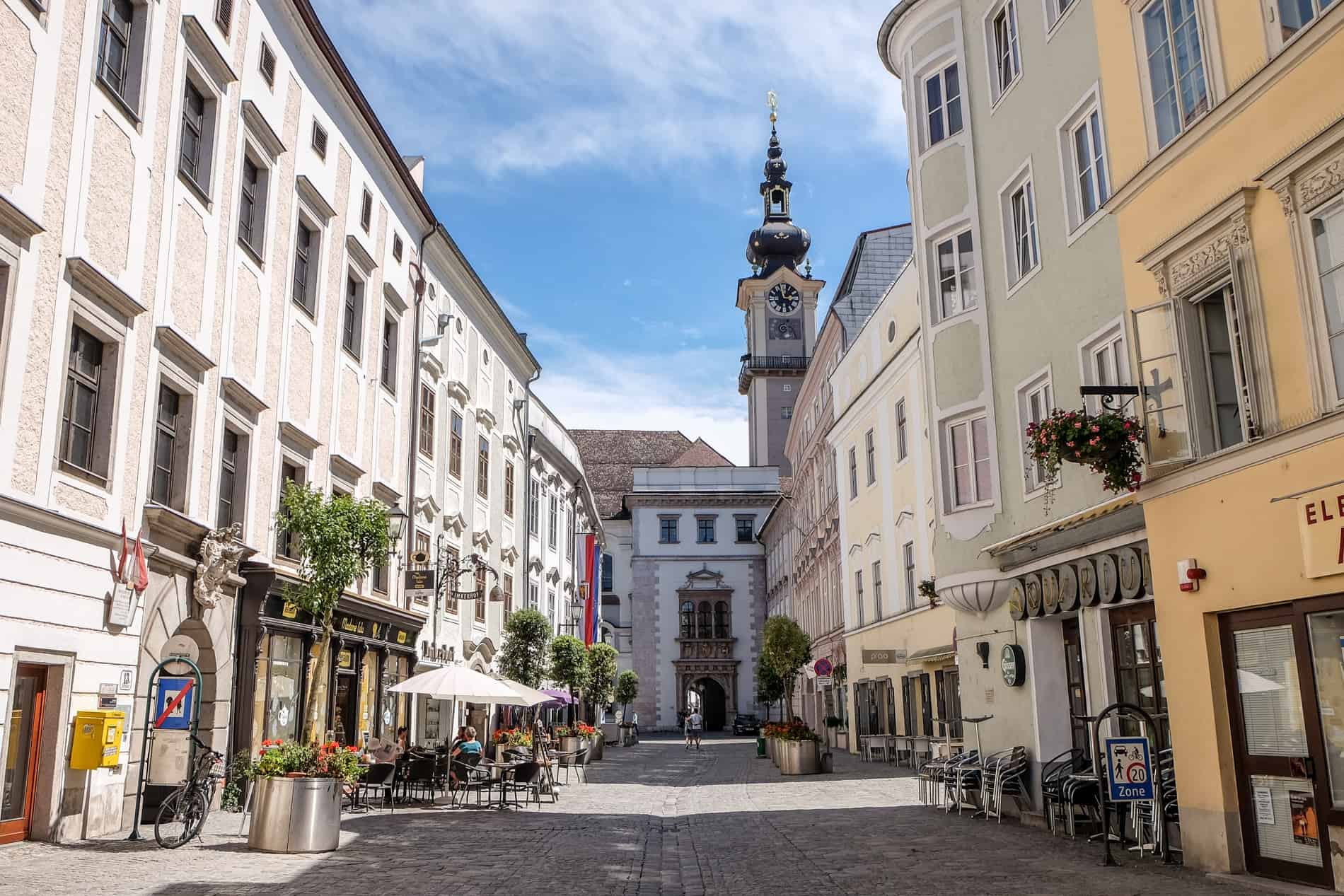 A shop and cafe lined street in Linz in Imperial style leading to a clock tower at the end. 
