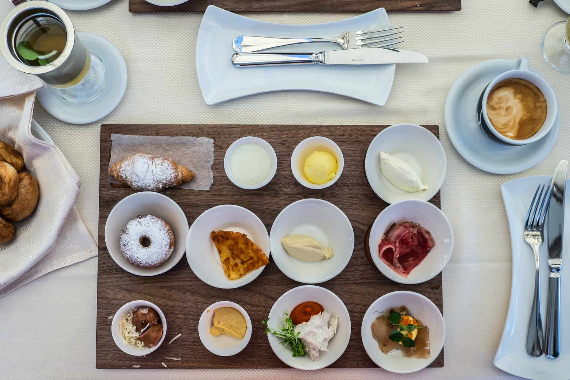 Elevenwhite cups full of various breakfast spreads, meats and cheeses presented on a wooden board with a croissant - part of the famous breakfast at Gostilna na Gradu at Ljubljana castle. 