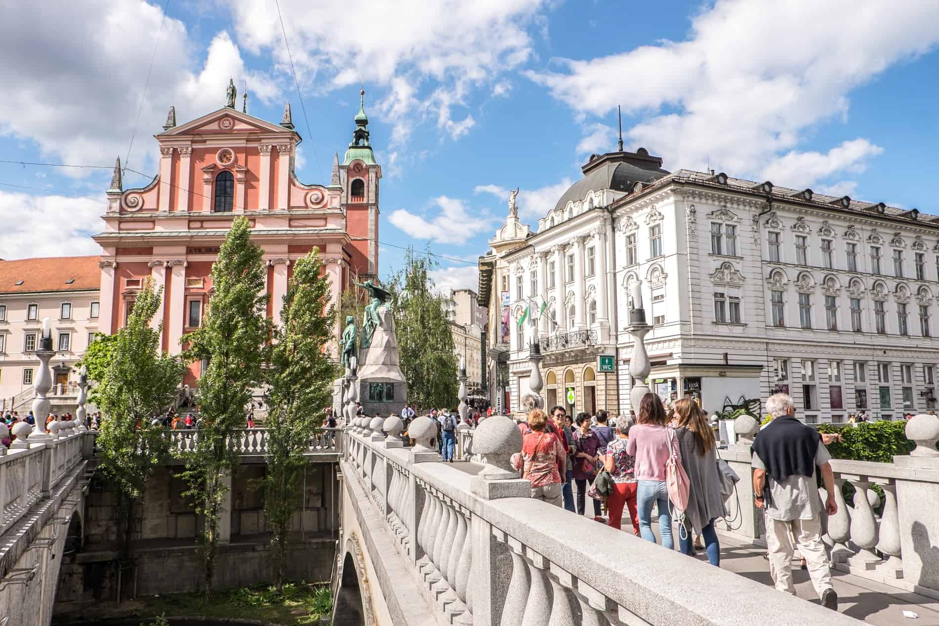 People walking over the Tromostovje Triple Bridge towards pinks and white classical buildings in Ljubljana, the Capital of Slovenia. 