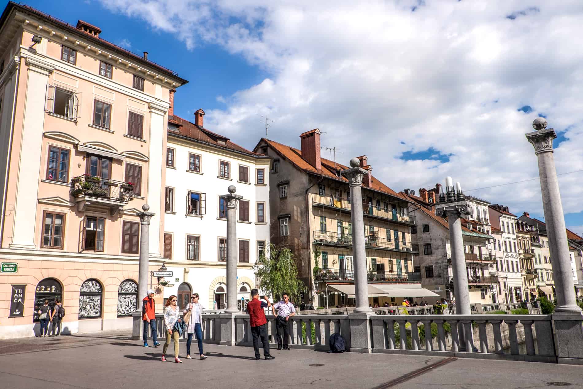 Poeple walking over the Plečnik designed Cobbler’s Bridge in Ljubljana in front of old town buildings. 