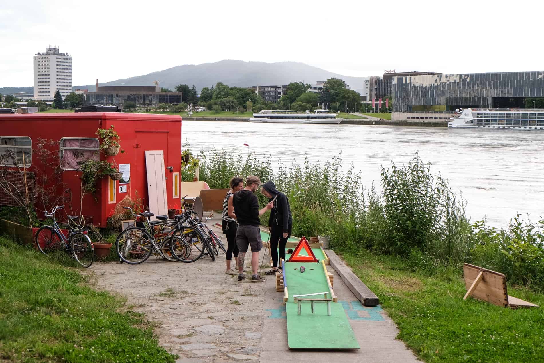 People standing between a red cafe van and green crazy golf game on the Danube river bank in Linz, Austria. 