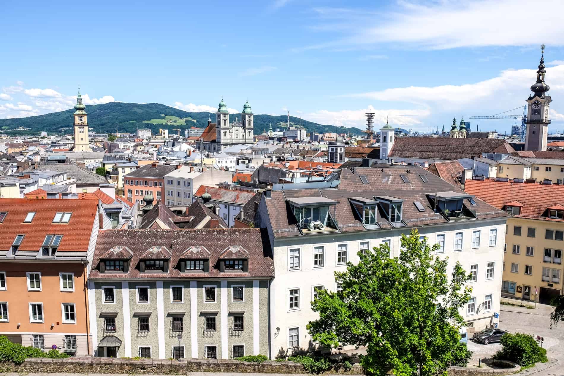 Elevated view over the city of Linz in Austria, with pastel buildings and church spires, backed by hills. 