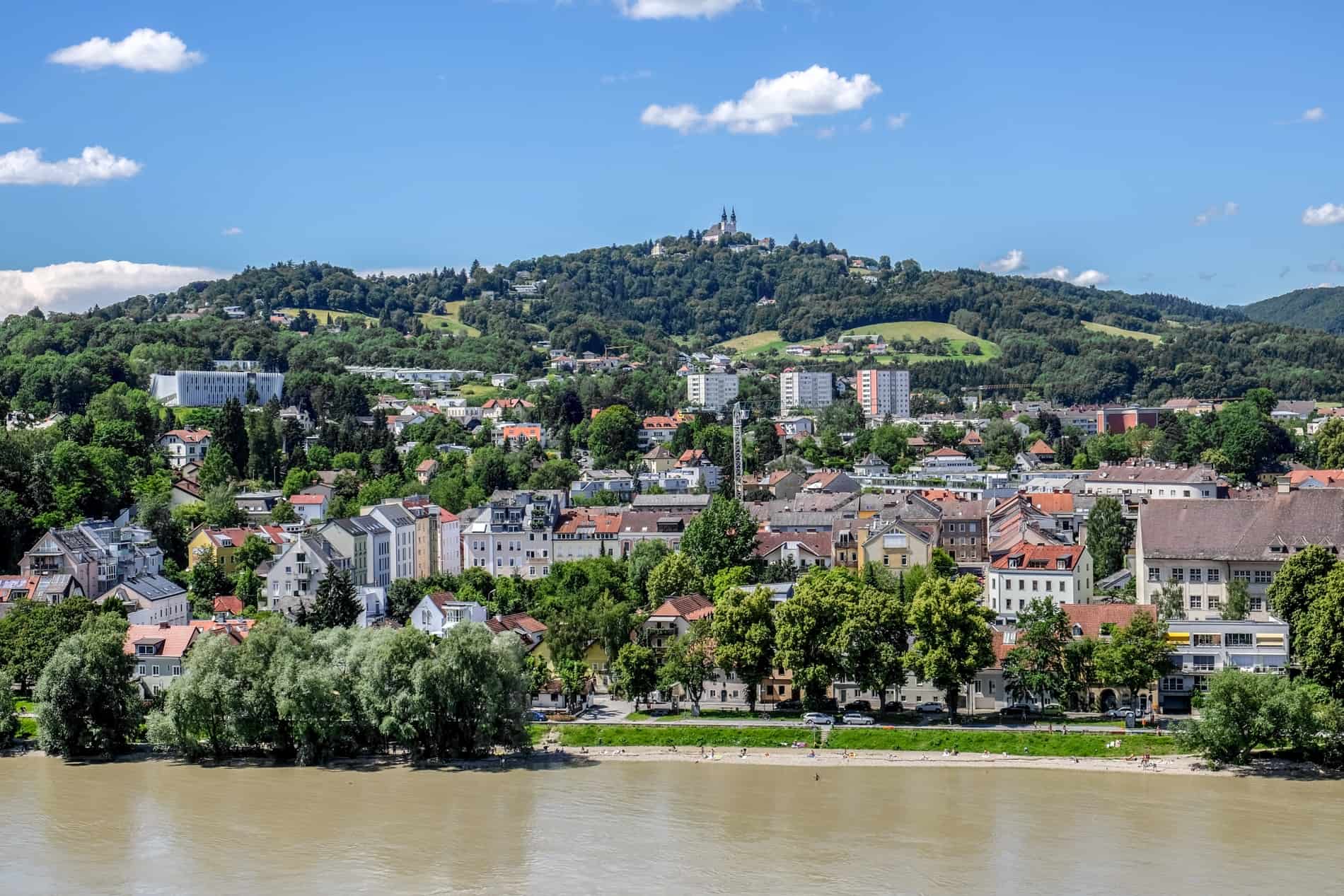 View across the rover in Linz to a green bank of pastel hued houses and a hilltop basilica. 