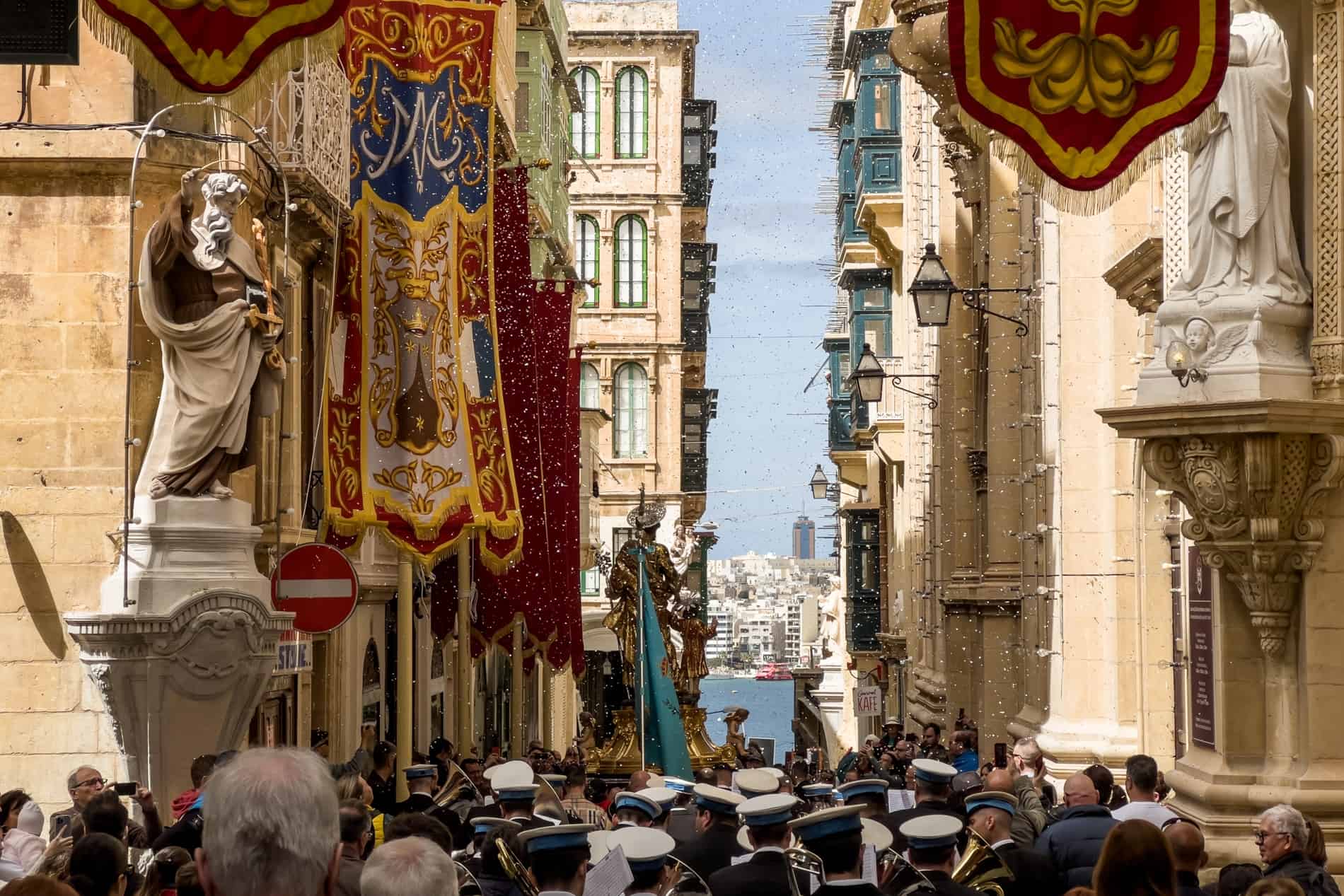 People follow a band performing in a street in Valletta, Malta. Red, gold and blue flags hang from the buildings. 