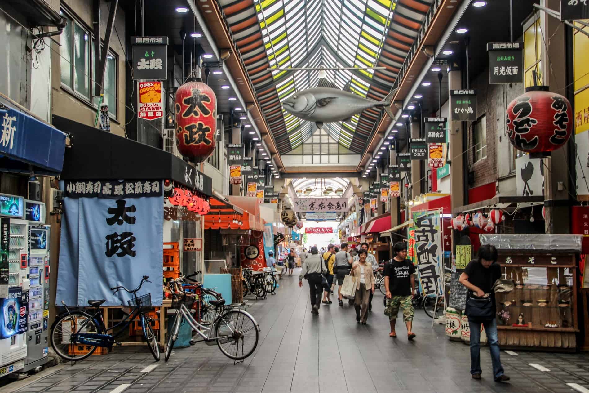 The covered street food arcade of Kuromon Ichiba Market in Osaka, lined with stalls and red lanterns. A giant fish sculpture hangs below the coloured glass ceiling. 