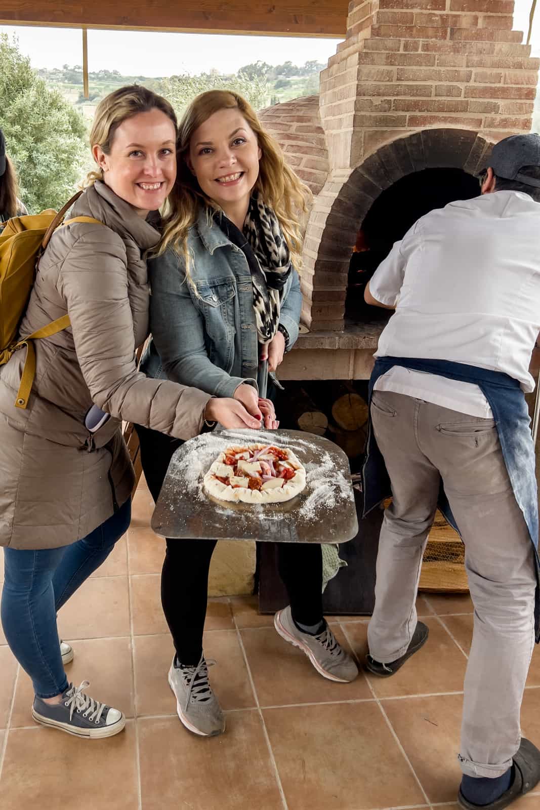 Two women holding a pizza ladle with a Maltese Ftira (sourdough bread and toppings) when a man fires up the wood oven next to them. 