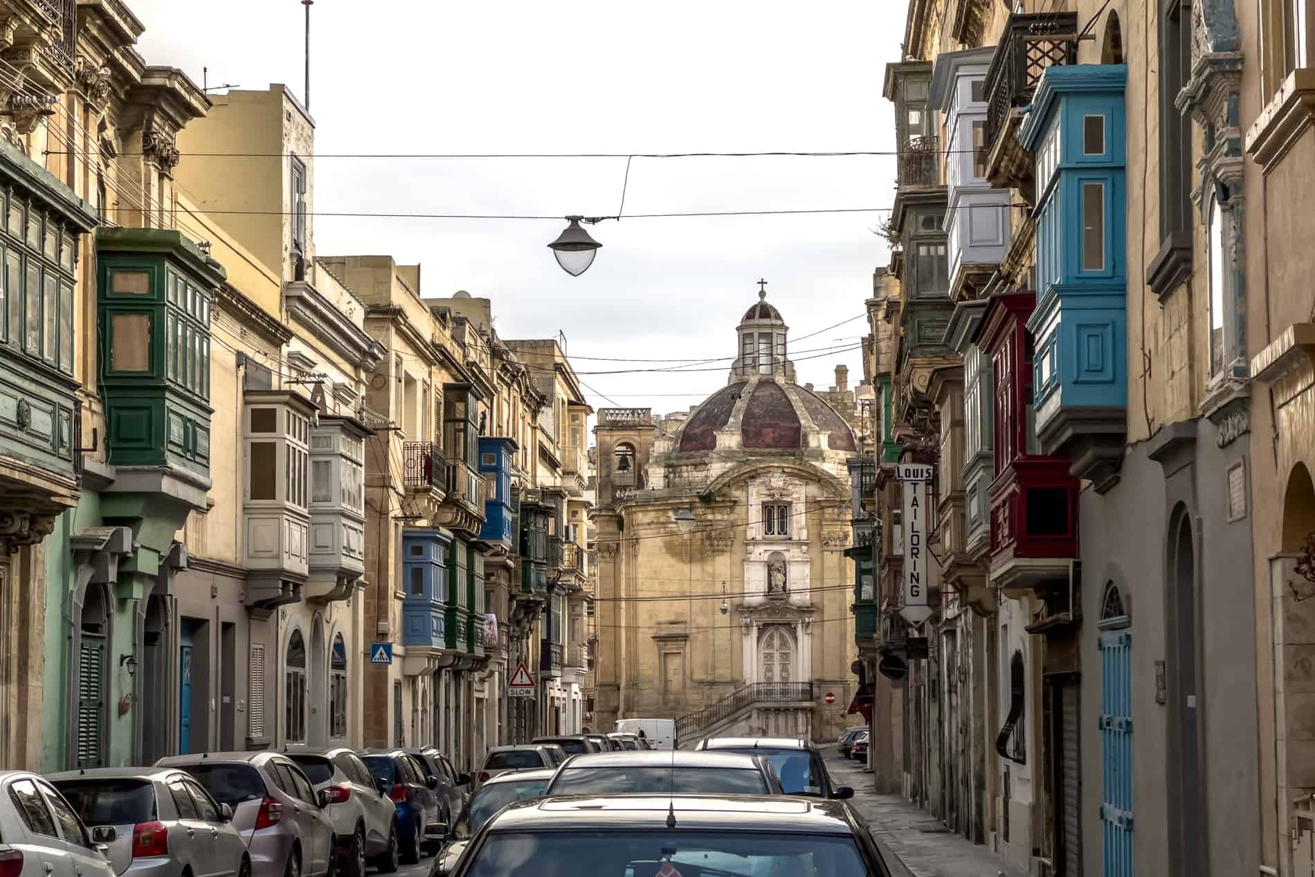 Honey-coloured buildings with blue, red, and green painted balconies on a street leading to a grand red-domed church building in Cospicua city, Malta. 