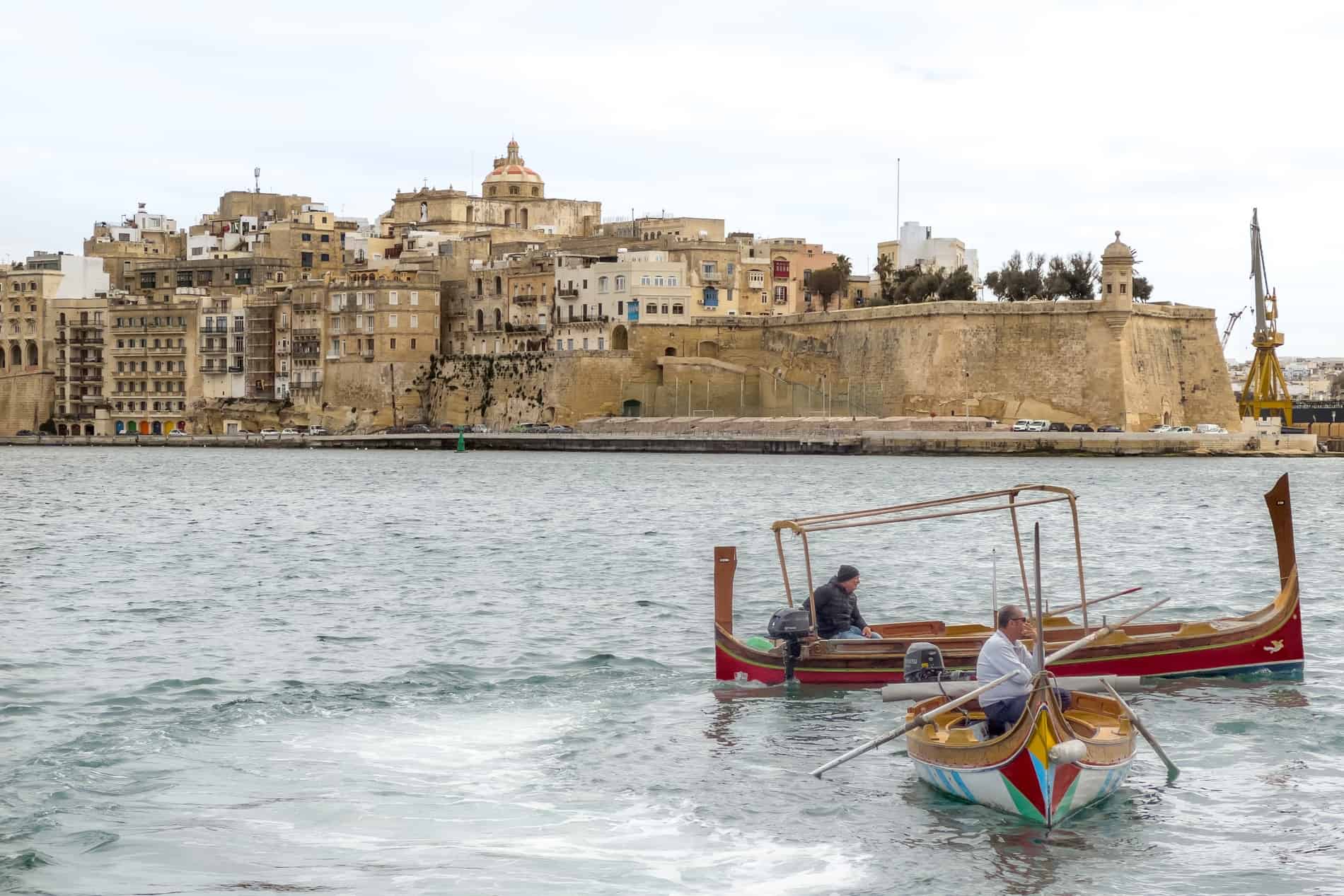 A peninsula of layered golden stone buildings and a fortification wall, jutting out into the waters of the Grand Harbour in Malta. 