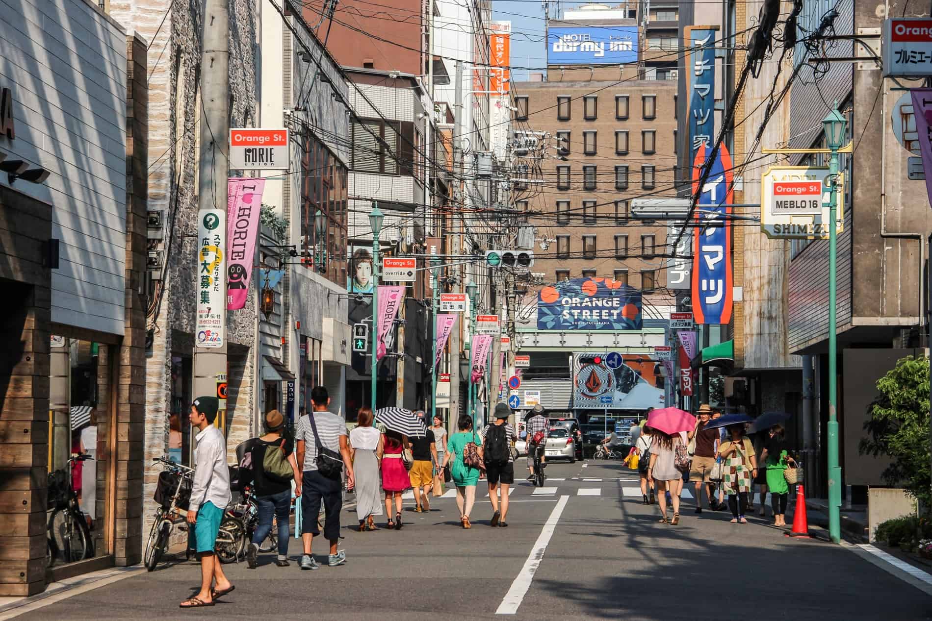 People walking on a wide street in Osaka lined with shop doorways. At the end of the street is a blue sign with orange dots that reads: Orange Street.