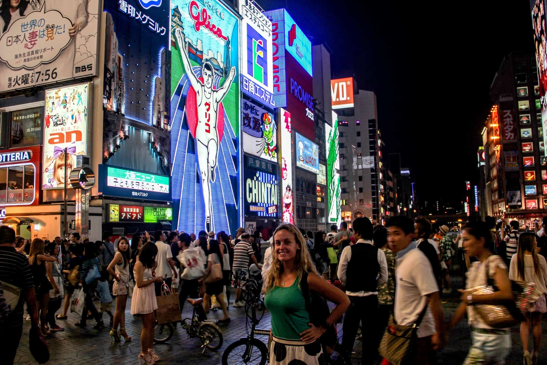 People gather in front of a building covered in animated advertising signs in Osaka, with the famous Glico Running Man sign in the middle. 