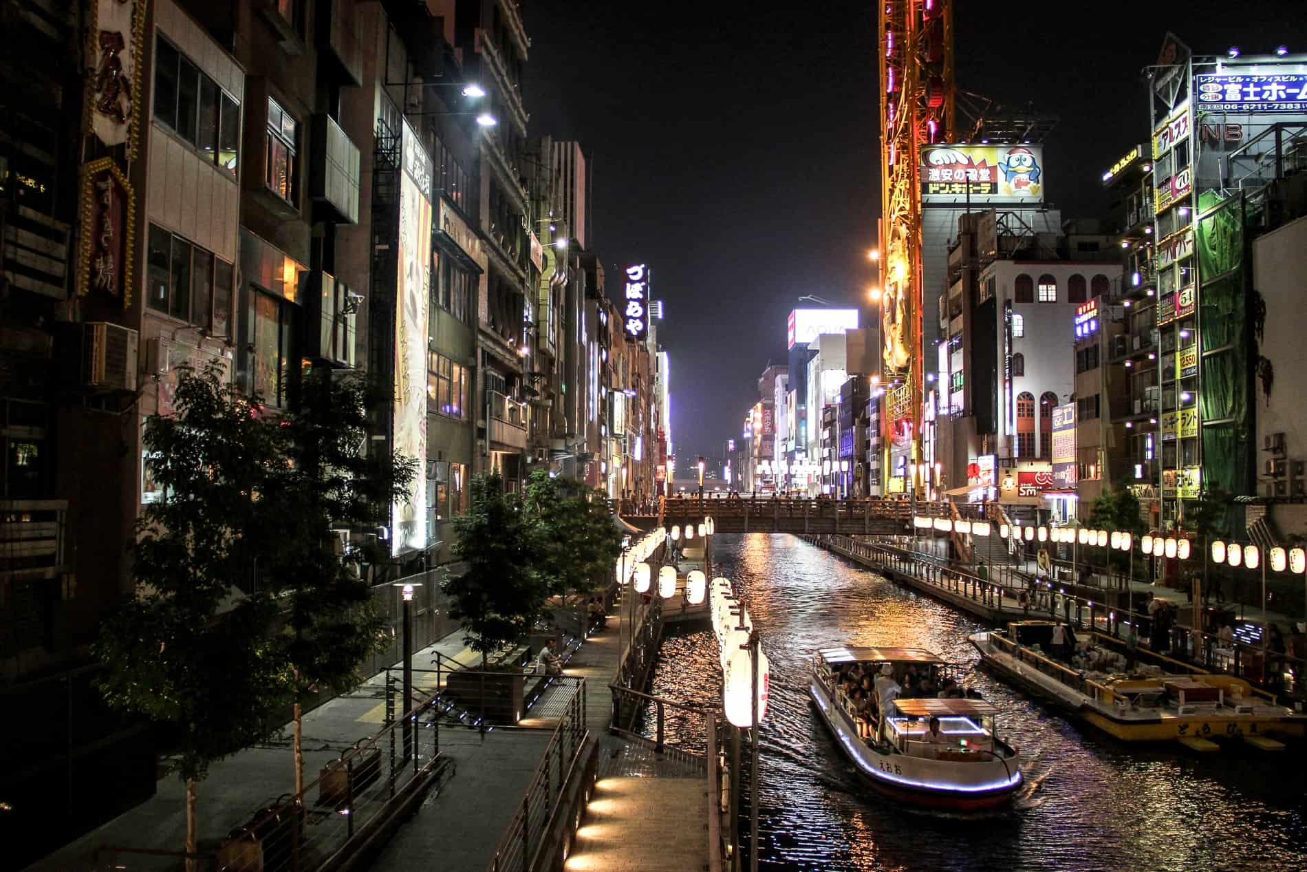 A small boat cruises down the river in Osaka on a canal lined with brightly lit buildings and pavements known as the Tombori River walk. 