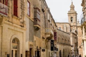 A row of honey-coloured limestone buildings in Mdina, Malta, with beautiful balconies, arched windows and towering spires.