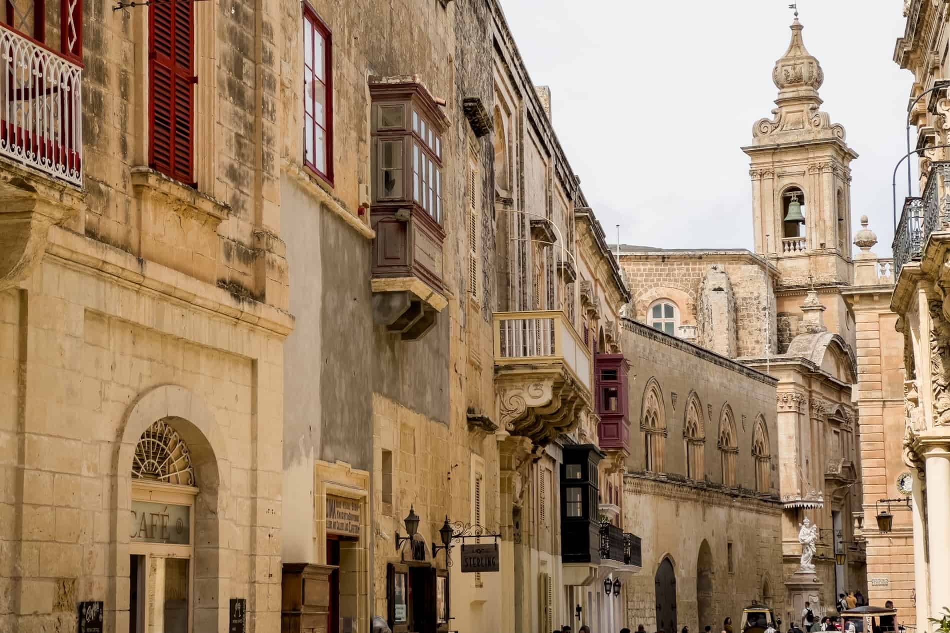 A row of honey-coloured limestone buildings in Mdina, Malta, with beautiful balconies, arched windows and towering spires. 