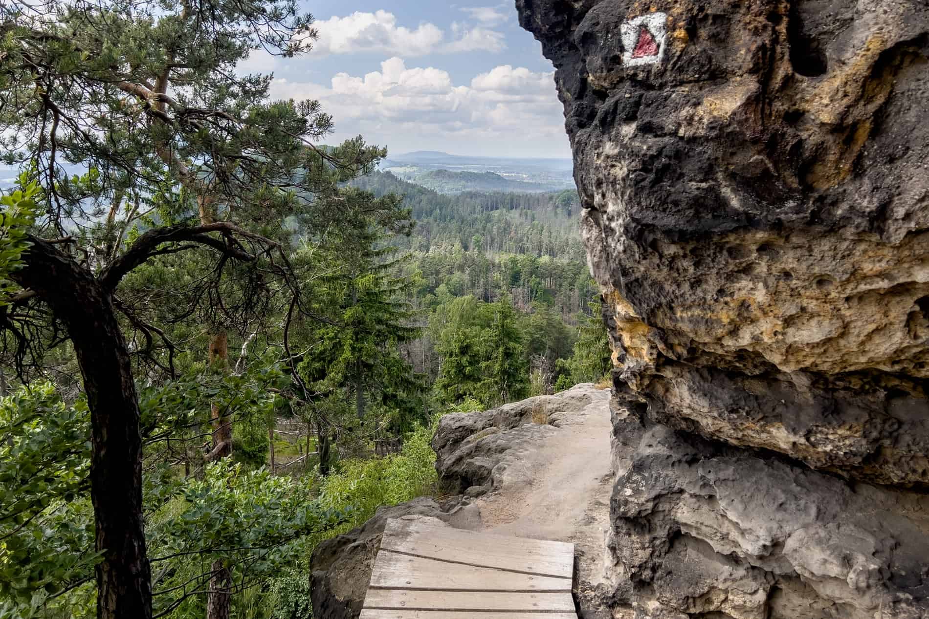 An elevated narrow footpath rounds a large rock with a view to Bohemian Switzerland National Park. 