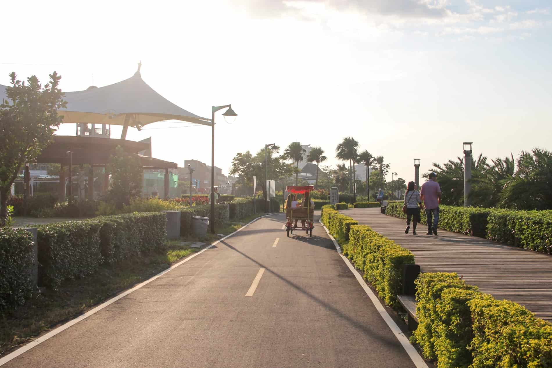 People walking on a wooden promenade and a tuktuk riding on the road in a green area of new Taipei. 