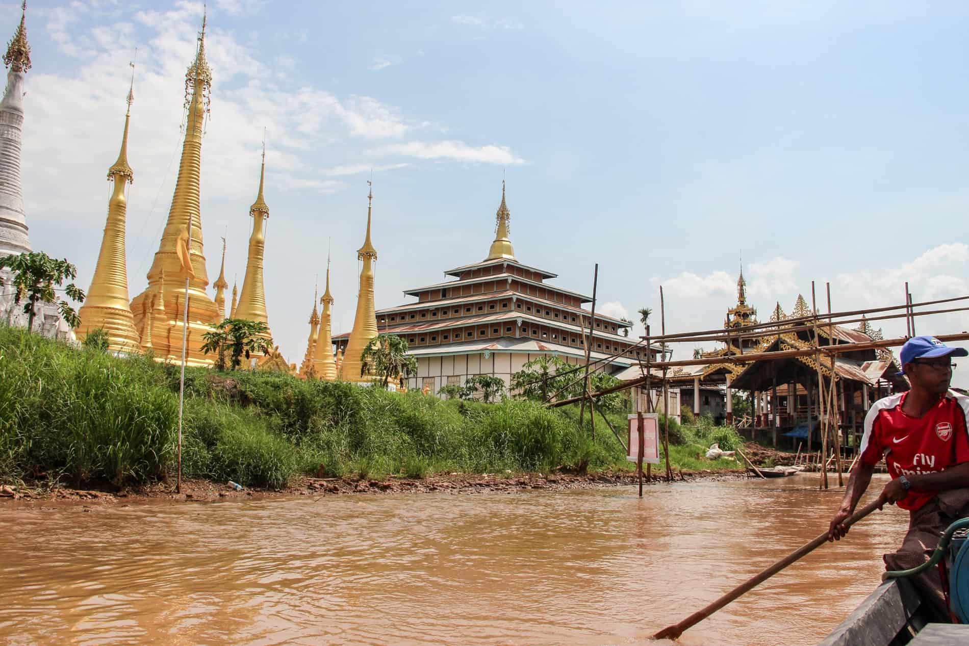 A man rows a boat past the golden stupas and tiered pagodas of a Buddhist Temple on Inle Lake. 