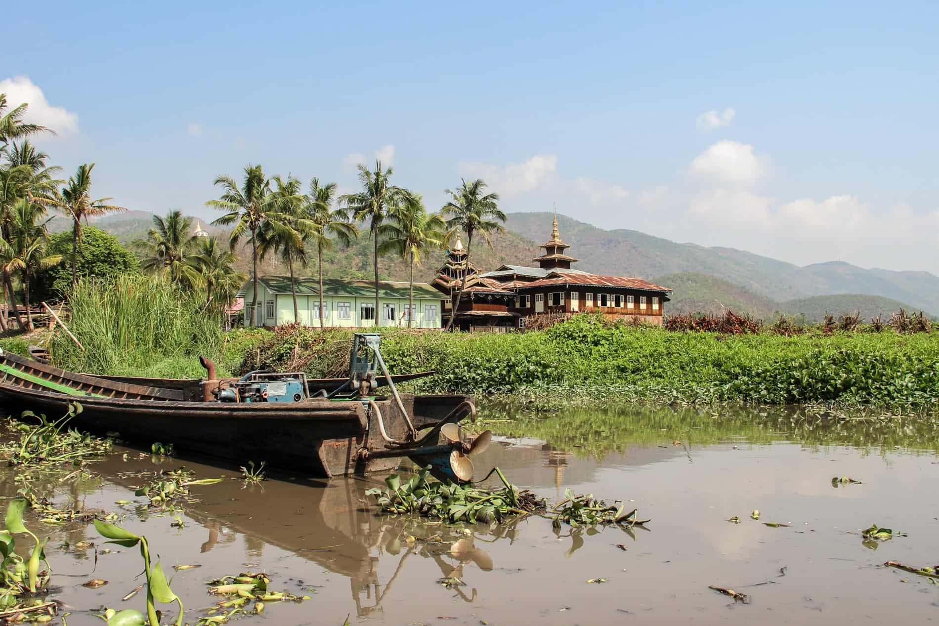 A rusting boat with a blue engine, sits in a pool of murky water, backed by green water plants. In the far distance is a red temple with a golden pagoda in the countryside of Myanmar, next to a mint green building and a row of six palm trees. 