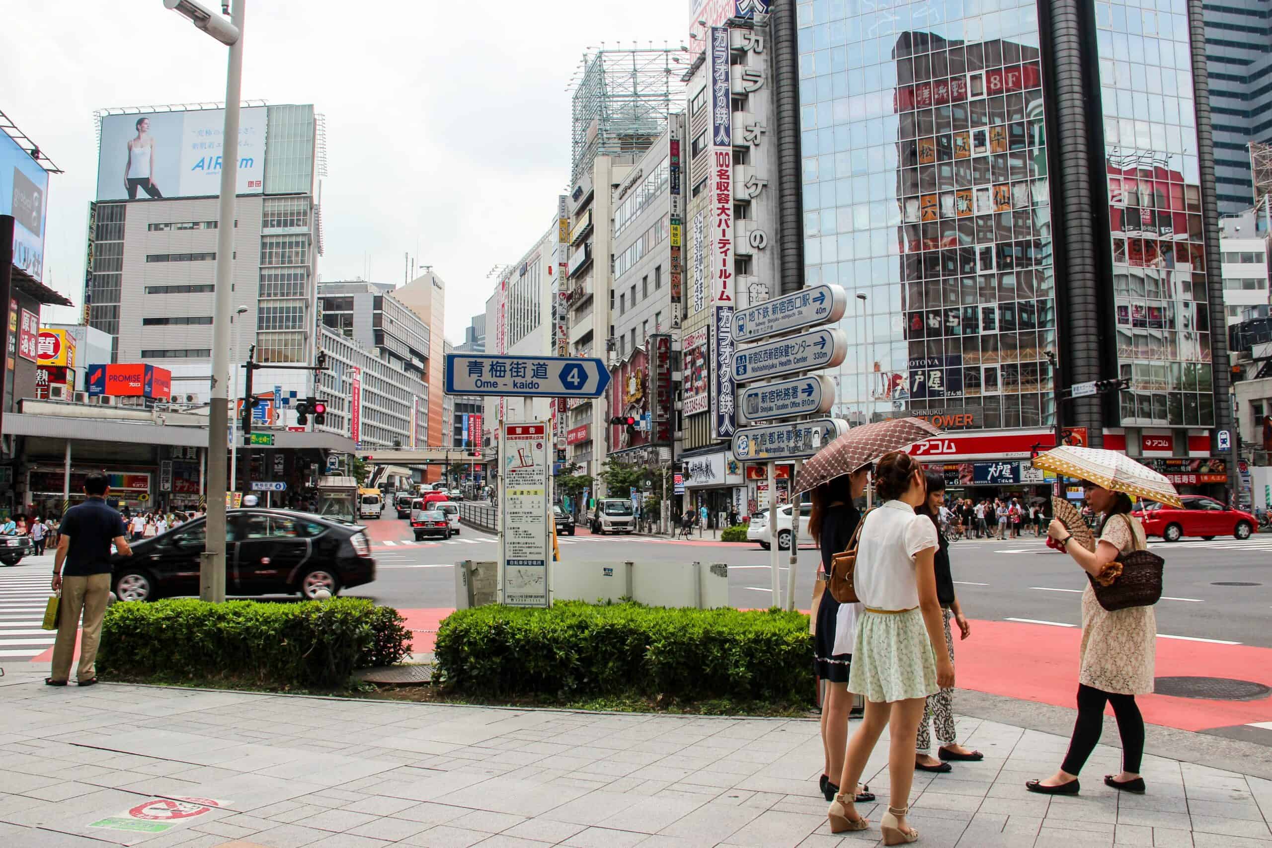 Four women on a street (two holding umbrellas) in modern Tokyo next to street signs.