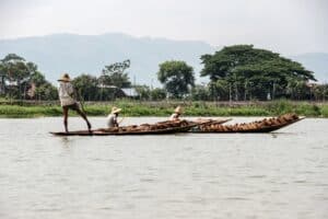 Three Inle Lake fisherman - one standing and rowing the oar with his leg and two seated - on boats stacked with wooden goods, in front of a floating garden.