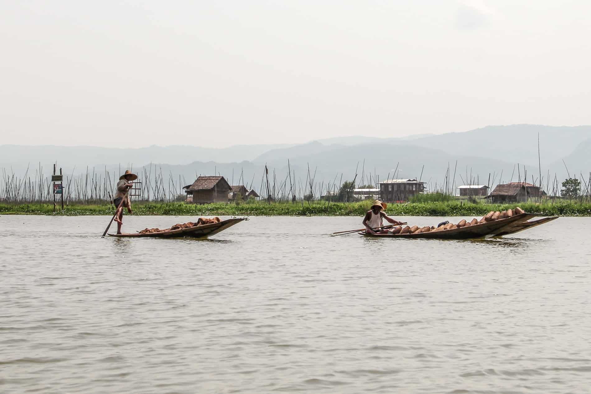 In front of a floating village in Inle Lake, a fisherman stands on the end of his boat rowing the oar with his leg. In front of him a man sits in his boat, heavy with solid objects. 