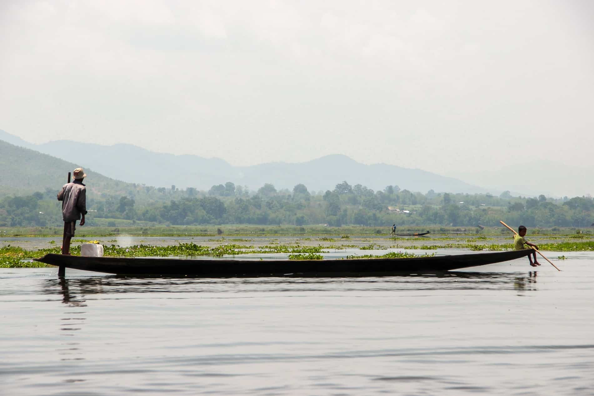 A standing man and a sitting young boy on the ends of a long wooden boat on the calm water of Inle Lake. 