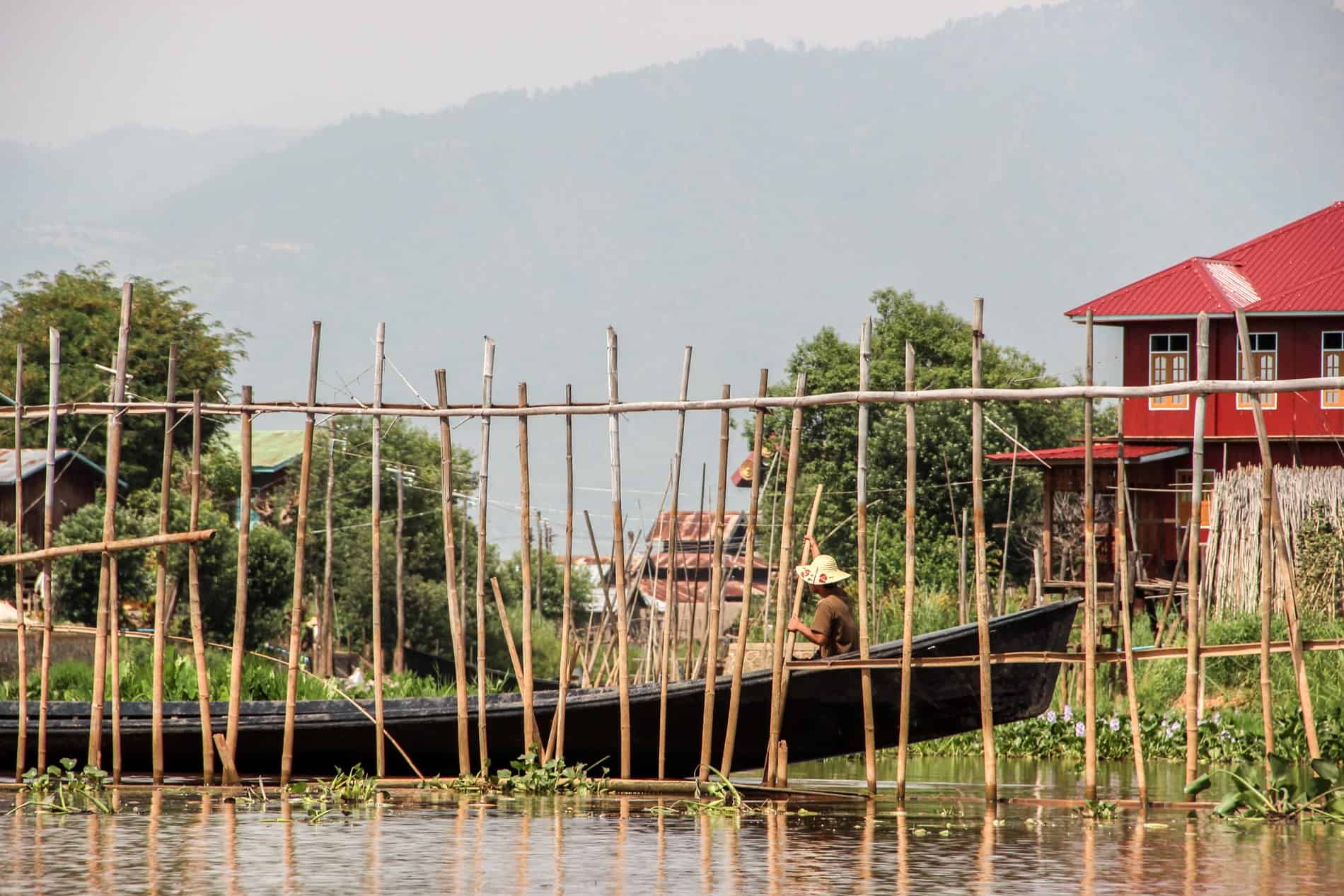 A person rows a long, dark wooden boat between a red houses and a bamboo pole wall in a floating garden on Inle Lake. 